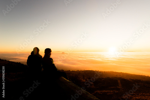 Silhouette of two friends man standing on a rock at sunrise on the fog of success and serenity.