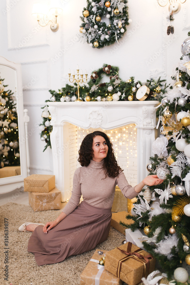 Attractive elegant beautiful brunette girl in a tight sweater and pleated skirt with a gift in her hands on the background of the fireplace, Christmas tree, garlands and bokeh in the white living room Attractive elegant beautiful brunette girl in a tight sweater and pleated skirt with a gift in her hands on the background of the fireplace, Christmas tree, garlands and bokeh in the white living room