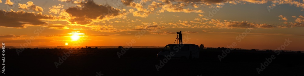 Panoramic of a field sunset with photographer taking pictures on top of the van