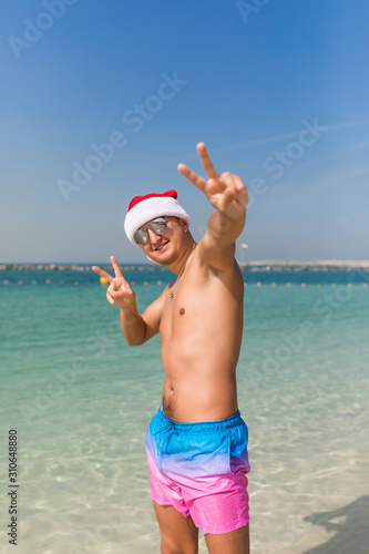 Young handsome man in santa hat and sunglasses with peace gesture standing in sea water background
