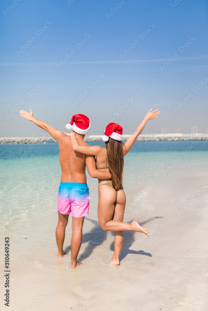 Back view happy couple in love walking on beach in santa hat. concept of celebrating the new year at sea