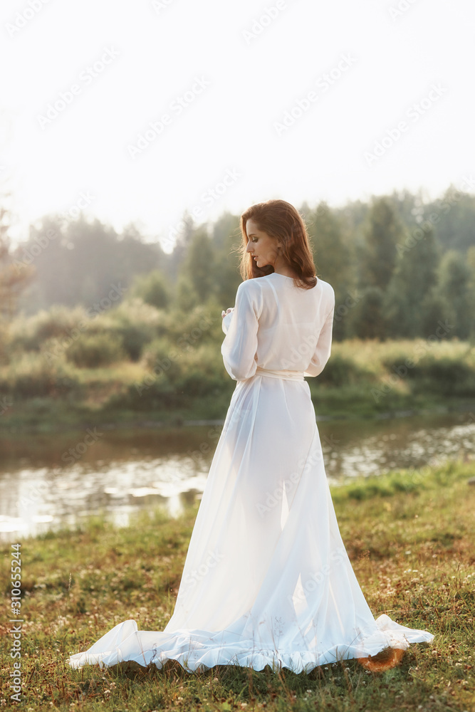 Beautiful slender woman in a long white dress walks in the morning near the lake. Brown-haired girl with long hair walks on the grass in the village, natural cosmetics and makeup