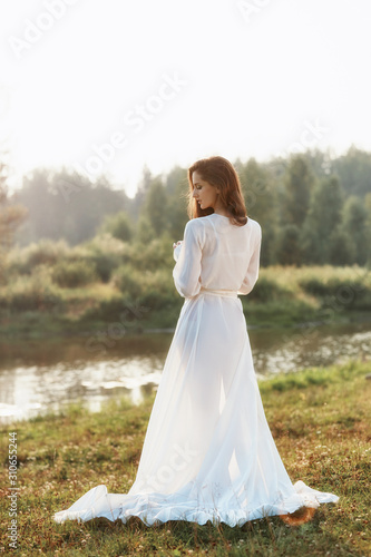 Beautiful slender woman in a long white dress walks in the morning near the lake. Brown-haired girl with long hair walks on the grass in the village, natural cosmetics and makeup