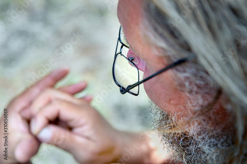 an old man with a beard and glasses looks at his hands. concept of old age and poor vision
