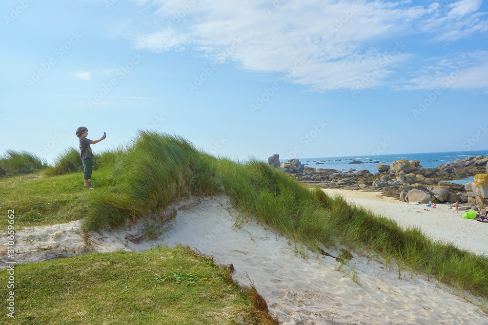 Obraz premium Interested boy taking a picture at a beach at a sunny and windy summer day, standing on a dune
