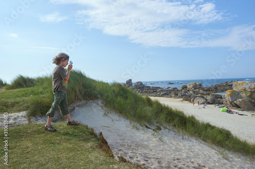 Interested boy concentrating on taking a picture, standing on a dune on a sunny and windy summer day on a beach in Brittany