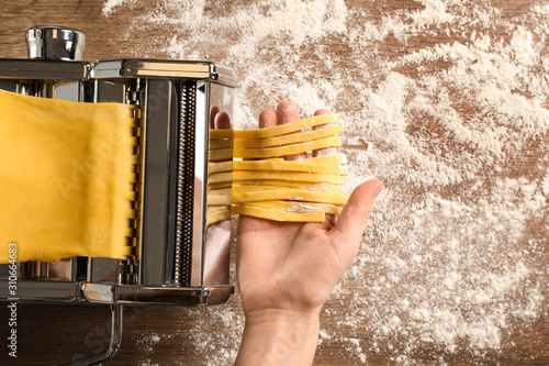 Woman preparing noodles with pasta maker machine at wooden table, top view