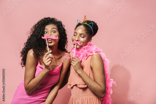 Image of cheerful african american women making fun with toy mustache