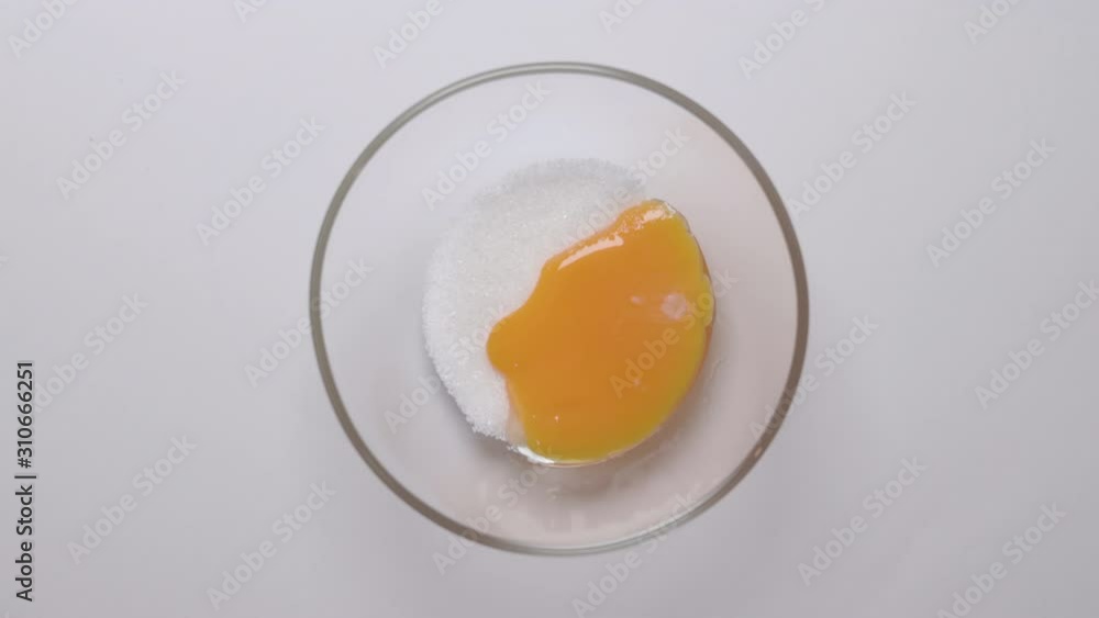 Top view from above of a glass bowl with sugar. Chicken egg yolk falls to the bowl from a spoon. Top view from above of a glass bowl with sugar. Chicken egg yolk falls to the bowl from a spoon.
