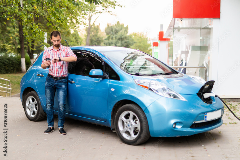 Young handsome man look at watch near his electric car charging at station Young handsome man look at watch near his electric car charging at station