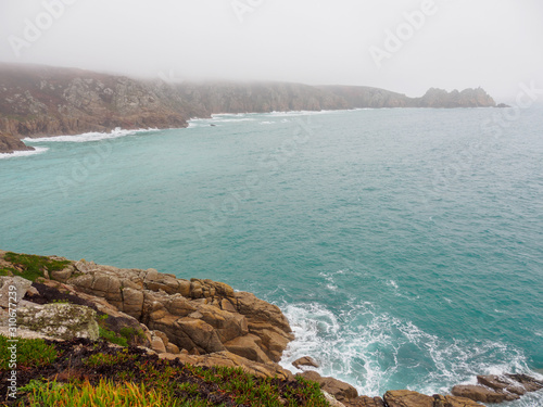 Wide angle view of the rocky shores and calm waters of Porthcuno beach on a foggy day. Cornwall, United Kingdom. Travel and nature. photo