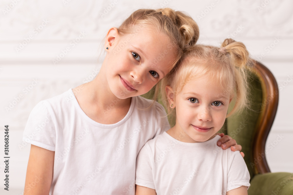 Two little blond smiling girls pose for camera while sit in green wooden armchair Two little blond smiling girls pose for camera while sit in green wooden armchair