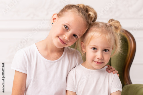 Two little blond smiling girls pose for camera while sit in green wooden armchair