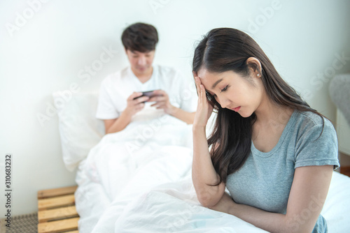 Young asian woman is upset that man uses phone all the time without paying attention to her. Man holds phone pay attention on smart mobile phone while sit on bed with ignoring on his girlfriend.