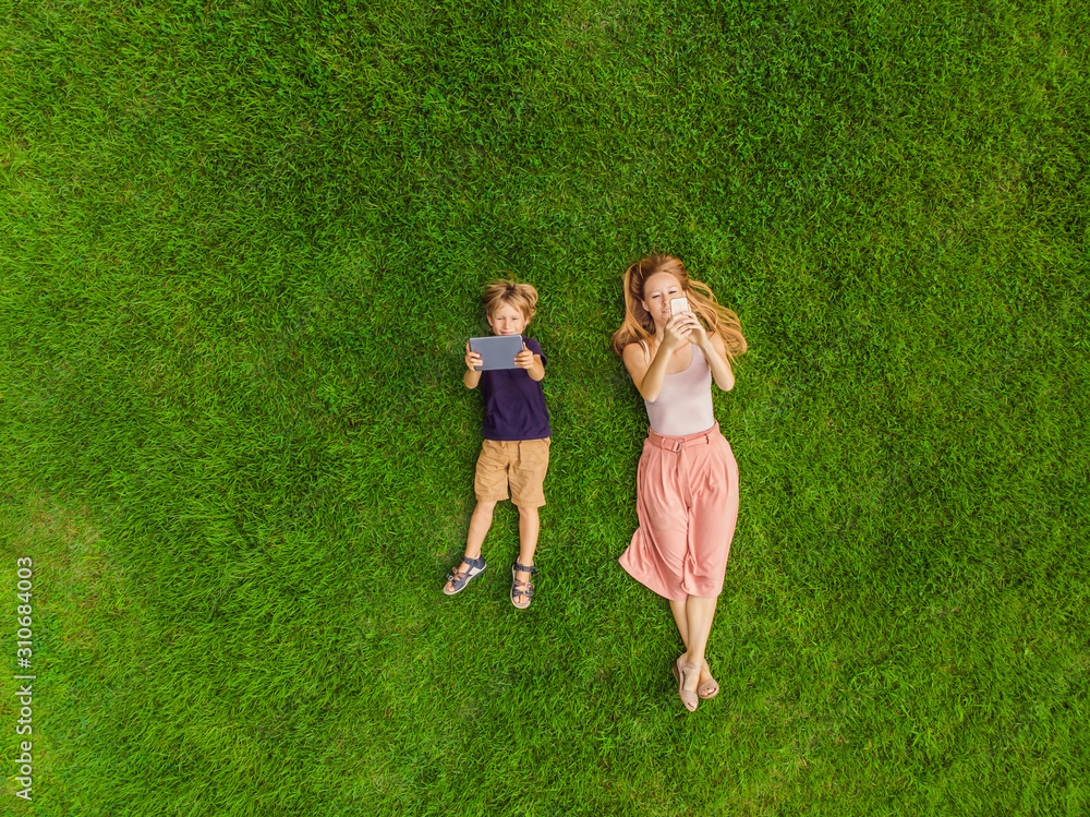 Obraz premium Mom and son are lying on the grass in the park. Mom looks at the phone, son looks at the tablet. Photos from the drone, quadracopter