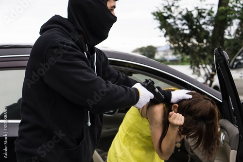 Dangerous criminal man with gun stealing car of scared young woman on outdoor parking.