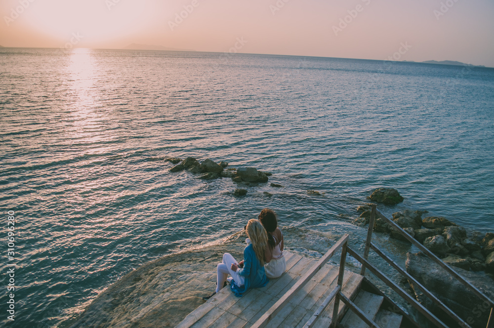 Two girls sitting and waiting for sunset. Blonde and brunette, friends traveling together. Corfu island, Greece. Two girls sitting and waiting for sunset. Blonde and brunette, friends traveling together. Corfu island, Greece.