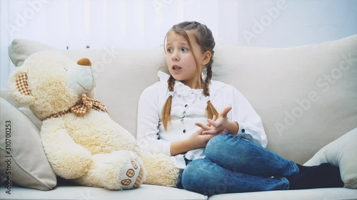 Cute little girl with plaits sitting on the sofa, talking with her teddy, bending her fingers desparately. photo