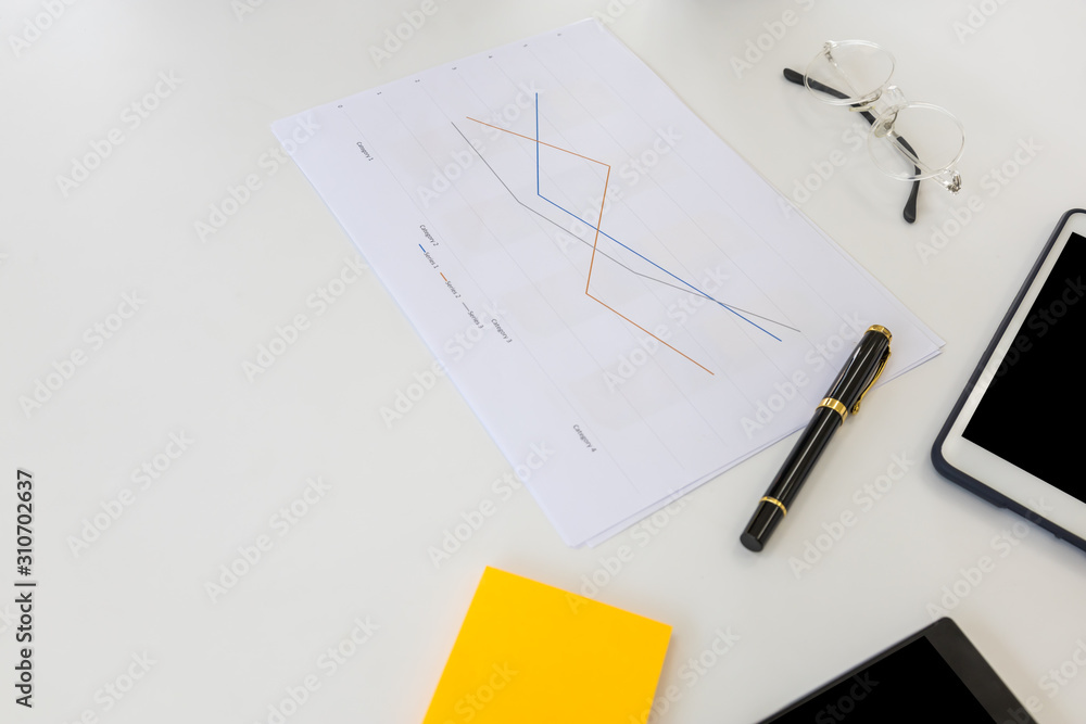 Business and Work Place Concept. Photo of line chart paper with pen, reading glasses, note, computer tablet and smart phone on white table. Business and Work Place Concept. Photo of line chart paper with pen, reading glasses, note, computer tablet and smart phone on white table.