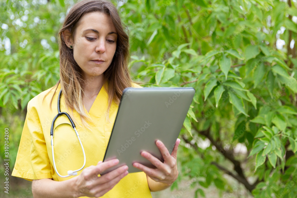 Mujer enfermera joven vestida con uniforme amarillo y estetoscopio colgado del cuello observando con atención la tablet que sostiene con ambas manos en entorno de naturaleza Mujer enfermera joven vestida con uniforme amarillo y estetoscopio colgado del cuello observando con atención la tablet que sostiene con ambas manos en entorno de naturaleza