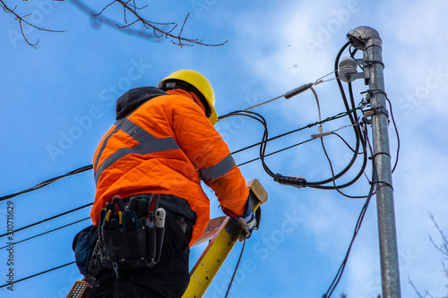 A telecoms operative is seen working from a ladder on a utility pole, wearing high visibility personal protective clothing, high viz PPE, and hard hat photo