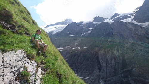 Very steep mountain meadow with emerald green grass exhausted hiker climber man boy is sitting on the edge enjoying the view and relaxing in the Swiss Alps Mountains and glaciers background