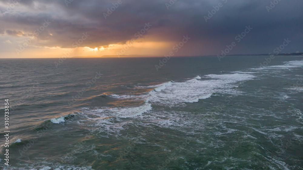 An aerial view of a choppy sea with crashing waves under a stormy cloudy grey sky with ray of light from the sun behind it