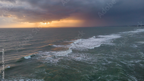 An aerial view of a choppy sea with crashing waves under a stormy cloudy grey sky with ray of light from the sun behind it © Dolwolfian