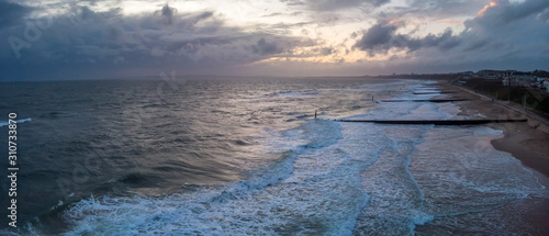 An aerial panoramic view of a choppy sea with crashing waves, groynes (breakwater), sandy beach and city in the background under a stormy cloudy grey sky