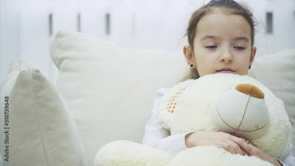 Small charming girl is sitting on the sofa and hugging her big plush friend, sighing. She seems upset and worried. Small charming girl is sitting on the sofa and hugging her big plush friend, sighing. She seems upset and worried.