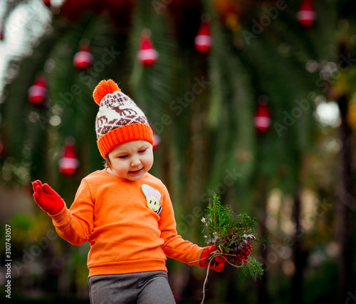 a small, beautiful girl in bright clothes,and in an orange hat, dancing near a Christmas tree, with a Christmas bouquet in her hand