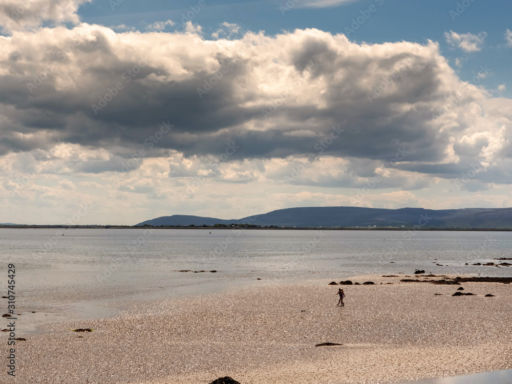 Silhouette of two woman on a sandy beach walking towards water, Galway bay, Burren mountains in the background, Cloudy sky over ocean.