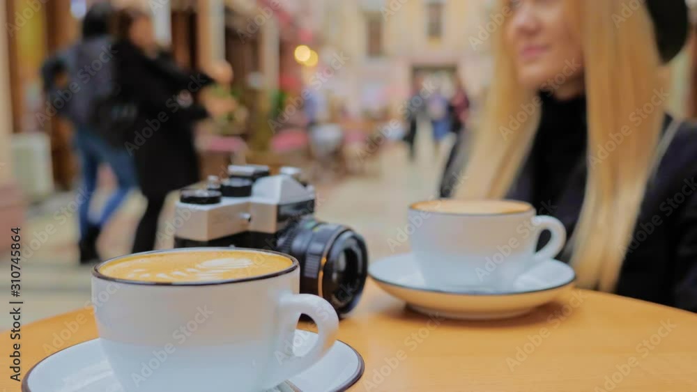 An unrecognized Caucasian woman, a blonde sitting at a table in a cafe. There are two cups of cappuccino, coffee and an old retro vintage film camera on the table. Unrecognized tourists and locals An unrecognized Caucasian woman, a blonde sitting at a table in a cafe. There are two cups of cappuccino, coffee and an old retro vintage film camera on the table. Unrecognized tourists and locals