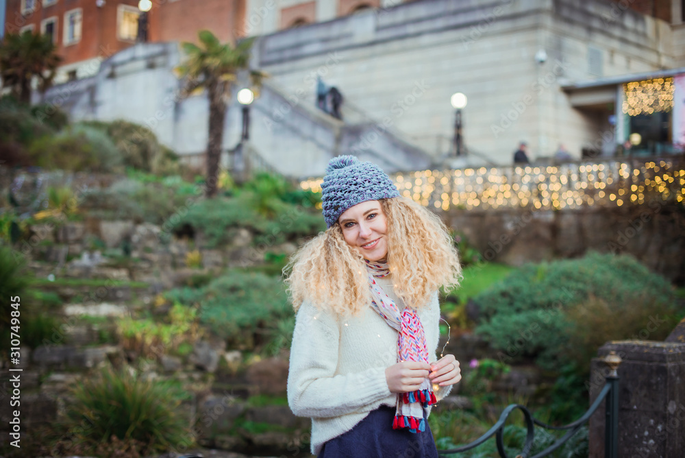 Portrait of smiling young curly blond woman in warm white natural sweater and blue knitted beanie hat with pompom looking at camera with the green park background and bokeh abstract lights.