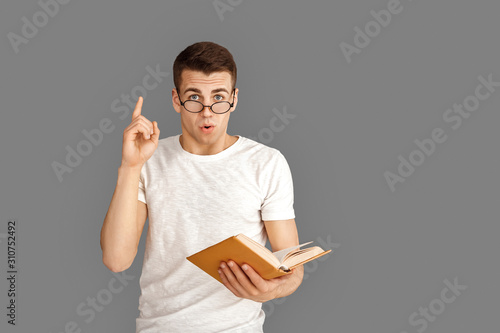 Freestyle. Young student in eyeglasses standing isolated on grey with book pointing up idea