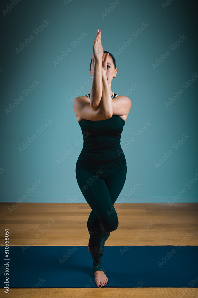 Sporty young woman doing stretching standing. Slim girl practicing yoga indoors on blue background. Calm, relax, healthy lifestyle concept.
