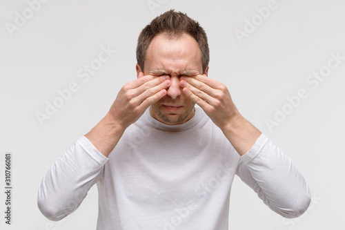 Close up of man has a problem with contact lenses, rubbing his swollen eyes due to pollen, dust allergy. Dry eye syndrome, watery, itching. Isolated on grey background, front view.  photo