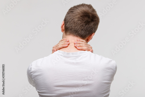 Back view of man in white top touching her pain in his neck and back, feels pain, massages the painful area. Cervical arthritis, osteochondrosis, diseases of musculoskeletal system