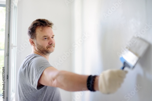 Portrait of a handsome Mature man making repairs in the apartment. The person paints the wall white. Do it yourself.