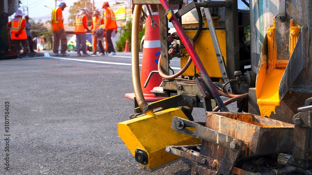 Fototapeta premium Close up thermoplastic spray marking machine with blurred background of road workers team discussing work on asphalt road in the city