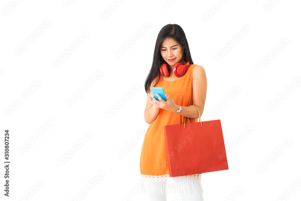 A beautiful Asian young woman wearing orange dress holding a red shopping bag using mobile phone with red head phone around her neck in front of white isolated background. A beautiful Asian young woman wearing orange dress holding a red shopping bag using mobile phone with red head phone around her neck in front of white isolated background.