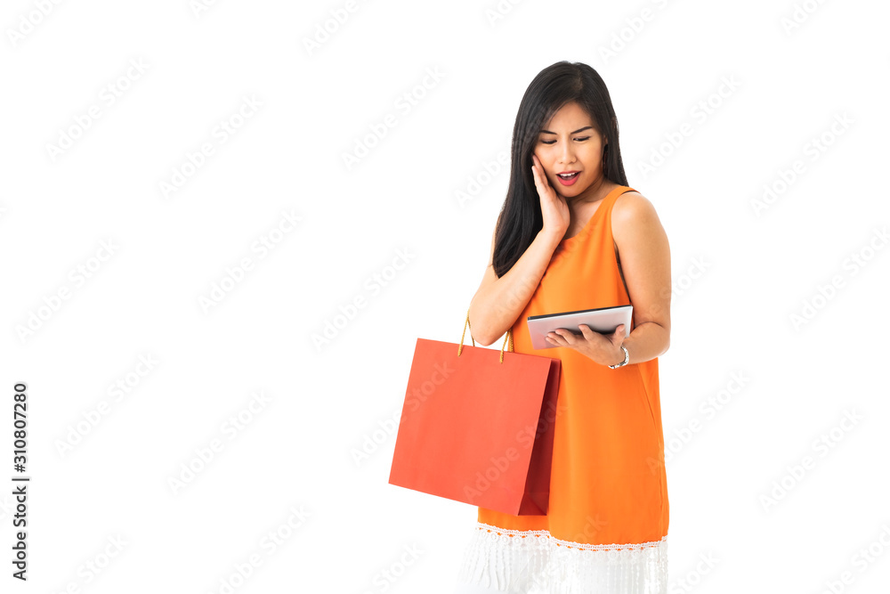 A beautiful Asian young woman wearing orange dress holding a tablet and a shopping bag looking surprise in front of white isolated background. A beautiful Asian young woman wearing orange dress holding a tablet and a shopping bag looking surprise in front of white isolated background.