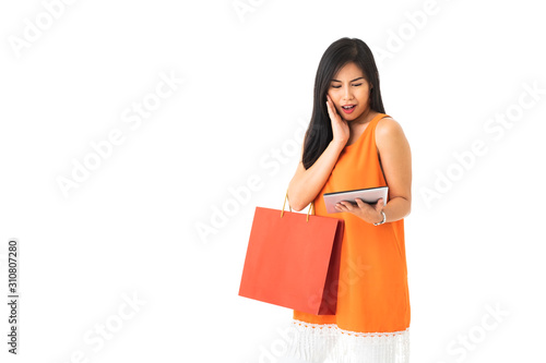 A beautiful Asian young woman wearing orange dress holding a tablet and a shopping bag looking surprise in front of white isolated background.