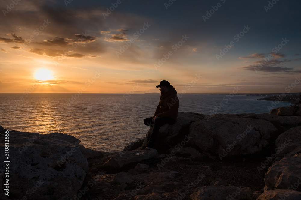Silhouette of a man sitting on a rock and watching the sunset in Cyprus, Cape Greco. Calm sea with orange sky. Copy space
