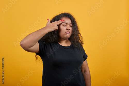 Afro-american woman with overweight keeping finger on head, like gun over isolated orange background wearing fashion black shirt. People lifestyle concepte.