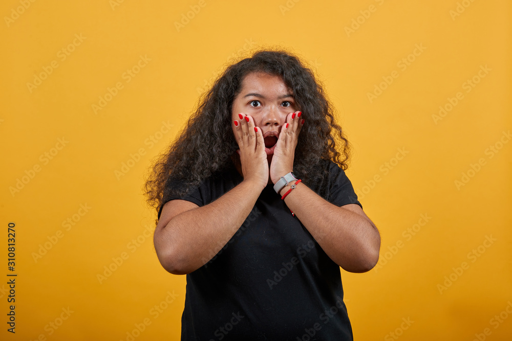 Shocked afro-american woman with overweight keeping hand on cheeks, looking at camera, confused over isolated orange background wearing fashion black shirt. People lifestyle concepte. Shocked afro-american woman with overweight keeping hand on cheeks, looking at camera, confused over isolated orange background wearing fashion black shirt. People lifestyle concepte.