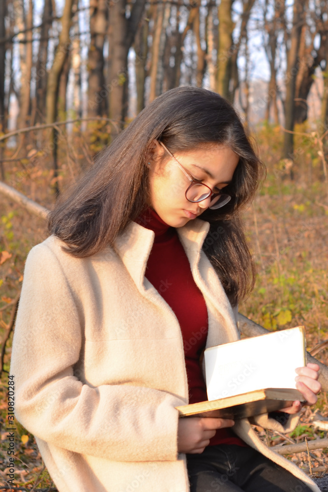 Obraz premium A girl in a light coat and glasses walks through the autumn Park with a book in her hands. The inscription on the book William Shakespeare. Soft focus.