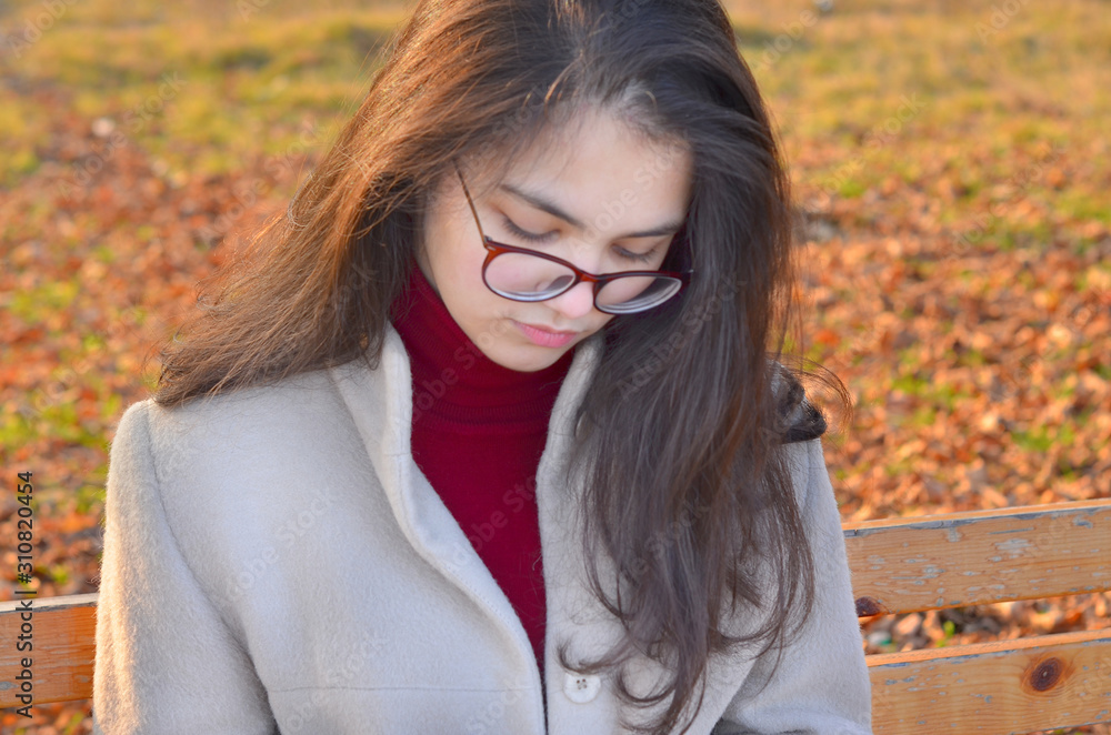 A young girl with dark hair in a light coat and Burgundy turtleneck sits on a bench in the Park and holds a paper Cup of coffee. Selective focus.