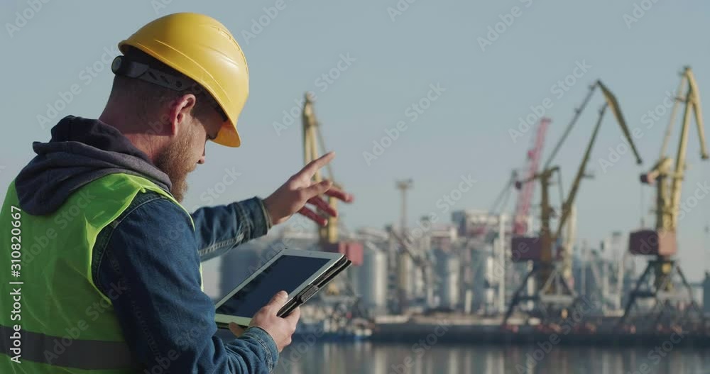 A Port Worker With A Beard In A Yellow Helmet, Works With A Tablet PC In The Seaport Against The Background Of Cranes. The Foreman Inspects The Industrial Harbor. Cinema 4K Video A Port Worker With A Beard In A Yellow Helmet, Works With A Tablet PC In The Seaport Against The Background Of Cranes. The Foreman Inspects The Industrial Harbor. Cinema 4K Video