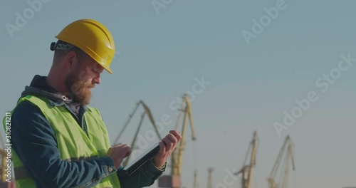Port Worker With A Beard In A Yellow Helmet Stands With A Tablet PC In The Seaport Against The Background Of Cranes. The Foreman Inspects The Industrial Harbor. Cinema 4K Video